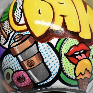 May include: A white ceramic sphere with a colorful, hand-painted design. The design features a yellow and brown word "DRINK", a red lip with a tongue sticking out, a green kiwi, a brown and silver coffee shaker, and a yellow and orange ice cream cone.