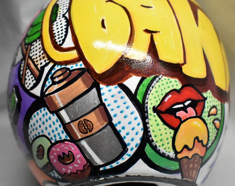 May include: A white ceramic sphere with a colorful, hand-painted design. The design features a yellow and brown word "DRINK", a red lip with a tongue sticking out, a green kiwi, a brown and silver coffee shaker, and a yellow and orange ice cream cone.