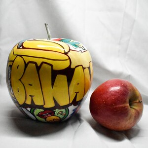 May include: A red apple and a yellow apple with the word "BANANA" painted on it in black and yellow letters.