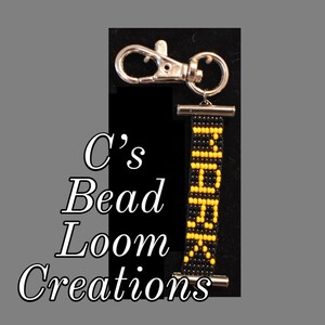 May include: Black and yellow beaded keychain with a silver clasp. The keychain is attached to a silver ring. The beads are arranged in a pattern that spells out the word "BEAD".