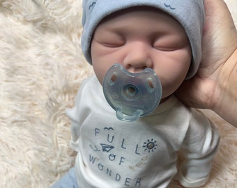 Puede incluir: Una muñeca bebé realista con gorro azul claro con diseños de olas, una camiseta blanca de manga larga con el texto "FULL OF WONDER" y pantalones azul claro. La muñeca tiene un chupete y está sobre una superficie blanca y esponjosa.