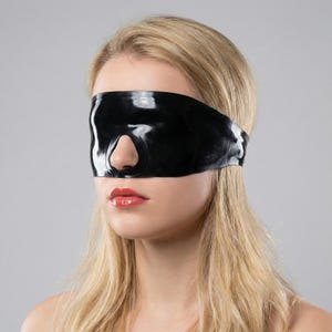 May include: A black, glossy blindfold with a nose cutout, worn by a person with blonde hair and red lipstick. The blindfold covers the eyes and forehead, creating a sleek, minimalist aesthetic. The background is a solid, neutral gray.