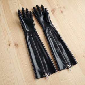 Pu&ograve; includere: Un paio di lunghi guanti neri con cerniere lungo le braccia. I guanti sono realizzati in un materiale lucido e sono appoggiati su una superficie di legno chiaro. I guanti hanno un design elegante e aderente.