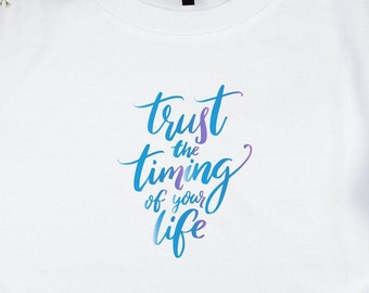May include: White t-shirt with the text "trust the timing of your life" in blue and purple script. The shirt is paired with light wash denim jeans, white sneakers, and black headphones on a wooden surface.