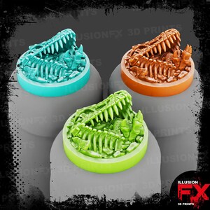 May include: Three circular miniature bases featuring a sculpted dinosaur skull and rib cage fossil design. One base is teal, one is orange, and one is lime green. Each base has a smooth, flat edge and a detailed, textured top surface.