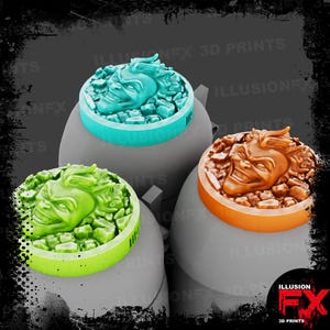 Puede incluir: Tres fichas de juego circulares con un rostro esculpido y sonriente que emerge de entre escombros. Una ficha es azul claro, otra verde brillante y otra naranja. Cada ficha tiene una base plana y lisa y una superficie superior texturizada.