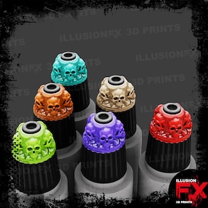 Könnte beinhalten: Eine Auswahl an dekorativen Aufsätzen in Schädelform, in den Farben Türkis, Orange, Beige, Grün, Lila und Rot. Sie sind auf schwarzen Sockeln montiert. Der Text "ILLUSION FX 3D PRINTS" ist im Bild enthalten.