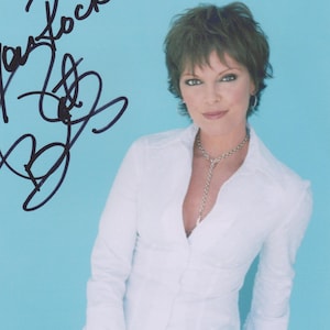 May include: A signed photograph of Pat Benatar. She is wearing a white button-down shirt and black trousers, with a silver necklace. The photo has the inscription "You Rock!" in black ink. The background is a light blue colour.