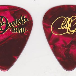 May include: Two guitar picks with a red and black marbled design. One pick features "The Charlie Daniels Band" in black and gold lettering. The other pick has a gold signature.