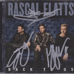 Può includere: Una copertina di CD con la band Rascal Flatts. La copertina ha uno sfondo blu con una foto in bianco e nero dei membri della band. I membri della band indossano tutti abiti casual. Il titolo dell'album è "Back To Us" ed è stampato in lettere bianche nella parte inferiore della copertina. La copertina del CD è firmata da tutti e tre i membri della band.