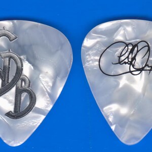 May include: Two pearlescent guitar picks. One pick features the initials "GB" in a stylized, dark gray font. The other pick displays a black signature. Both picks have a pearlescent, iridescent finish and are set against a blue background.