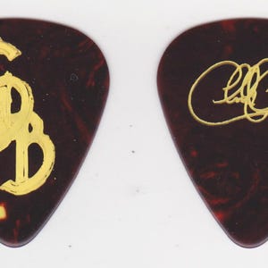 May include: Two guitar picks with a dark brown and red marbled pattern. One pick features the gold letters "GSB", the other has a gold signature. These picks are likely music memorabilia.
