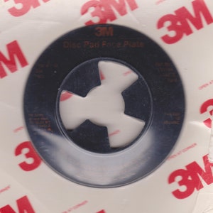 Puede incluir: Una placa frontal de almohadilla de disco de plástico negro con un logotipo de 3M y el texto "Disc Pad Face Plate".