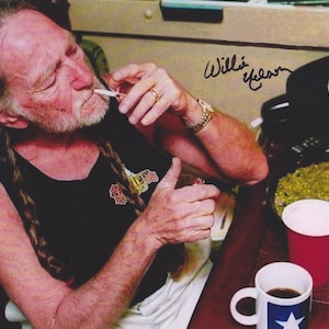 May include: An image of a person lighting a cigarette. The person is wearing a black tank top. A red cup and a mug with a blue star are on the table. The signature "Willie Nelson" is visible.