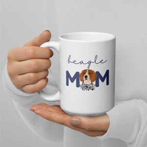 May include: White ceramic mug with the words "beagle MOM" in blue script and block letters. The letter "O" in "MOM" features a beagle illustration with a floral arrangement. The mug has a large handle.