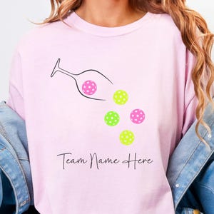 May include: A light pink t-shirt with a graphic of a wine glass pouring out pickleballs. The pickleballs are pink and green with white polka dots. The text "Team Name Here" is below the graphic. The word "BLOSSOM" is at the bottom.