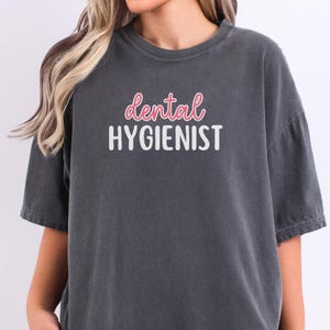 May include: A dark gray t-shirt with the words "dental HYGIENIST" printed on the front in white and pink lettering. The shirt has short sleeves and a relaxed fit. The word "PEPPER" is printed at the bottom.