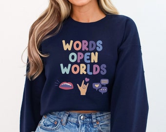 Puede incluir: Una sudadera azul marino recortada con el texto "WORDS OPEN WORLDS" en colores arcoíris. La sudadera tiene una variedad de iconos coloridos que incluyen una boca, una mano haciendo un signo de paz, un corazón y burbujas de diálogo.