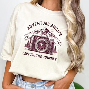 May include: Ivory-colored t-shirt with a maroon graphic featuring a vintage camera, mountains, and the text "Adventure Awaits, Capture the Journey." The design includes the numbers "19" and "17" on either side of the camera.