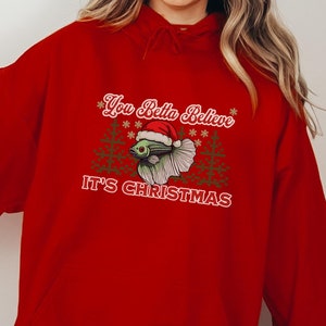 May include: Red hooded sweatshirt with a Christmas-themed design featuring a betta fish wearing a Santa hat and the text "You Betta Believe It's Christmas".
