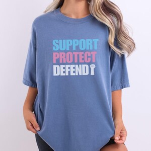 Może przedstawiać: Niebieska koszulka z napisem "SUPPORT PROTECT DEFEND" w kolorze różowym, niebieskim i białym z białym symbolem pięści.
