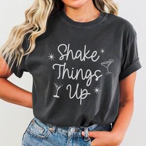 Könnte beinhalten: Dunkelgraues T-Shirt mit dem Schriftzug "Shake Things Up" in weißer Schrift, zusammen mit Cocktailglas-Illustrationen. Das Shirt hat eine lockere Passform und hochgekrempelte Ärmel. Das Wort "PEPPER" ist unten aufgedruckt.