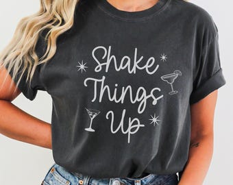 May include: Dark gray t-shirt with the phrase "Shake Things Up" in white script, along with cocktail glass illustrations. The shirt has a relaxed fit and rolled-up sleeves. The word "PEPPER" is printed at the bottom.