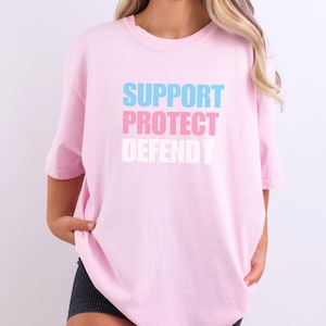 Może przedstawiać: Różowy T-shirt z napisem "SUPPORT PROTECT DEFEND" w kolorze niebieskim, różowym i białym.