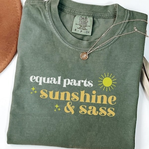 May include: A moss green t-shirt with the text "equal parts sunshine & sass" in white and yellow. The shirt has a crew neck and short sleeves. A gold necklace with a pendant rests on the shirt. The shirt is folded on a white surface.