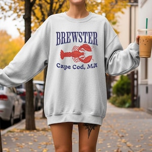 Könnte beinhalten: Hellgraues Sweatshirt mit dem Wort "BREWSTER" in Marineblau über einer roten Hummer-Grafik. Unter dem Hummer steht "Cape Cod, MA" in Marineblau. Der Pullover hat einen lässigen Stil.