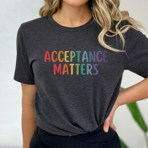 Könnte beinhalten: Ein dunkelgraues T-Shirt mit dem Text "ACCEPTANCE MATTERS" in Regenbogenfarben.