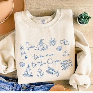 May include: A cream-colored crewneck sweatshirt featuring blue line art illustrations of a lighthouse, sailboat, seagull, anchor, lobster, fish, and nautical symbols, with the text "take me to the Cape" printed in the center in a cursive font.