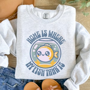 May include: Pale grey sweatshirt with the text "HOME IS WHERE MY FISH TANK IS" in a retro font. The graphic features a cartoon goldfish in a fishbowl with a light blue and yellow colour scheme. The sweatshirt is folded on a denim surface.