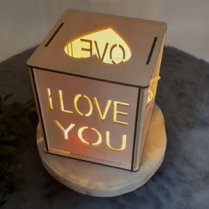 Könnte beinhalten: Eine weiße Holzkiste mit einem herzförmigen Ausschnitt oben und den Worten "I LOVE YOU" ausgeschnitten auf der Vorderseite. Die Kiste ist von innen beleuchtet und erzeugt ein warmes Leuchten.