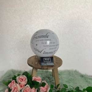 Può includere: Sfera di vetro bianca con un'iscrizione nera che recita "Freunde sind wie Sterne... Du kannst sie nicht immer sehen aber sie sind immer da". La sfera è appoggiata su uno sgabello di legno con un biglietto da visita sopra.