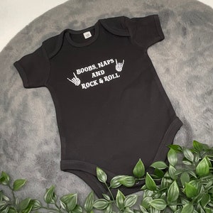 May include: Black baby onesie with white text that reads "Boobs, Naps and Rock & Roll" with two white skeleton hands making the rock and roll hand gesture.
