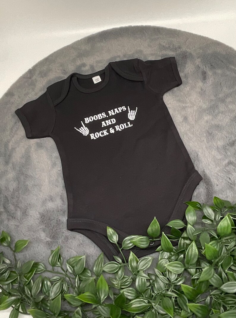 May include: Black baby onesie with white text that reads "Boobs, Naps and Rock & Roll" with two white skeleton hands making the rock and roll hand gesture.