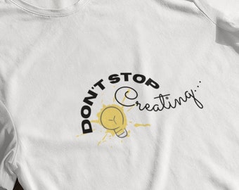 Può includere: Maglietta bianca con una grafica nera che dice "Don't Stop Creating..." con un'illustrazione di lampadina gialla.