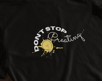 Può includere: Maglietta nera con una grafica bianca che dice "Don't Stop Creating" con uno spruzzo di vernice gialla e un orologio all'interno dello spruzzo.