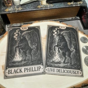May include: Two tarot cards featuring a black goat illustration with the text "BLACK PHILLIP" and "LIVE DELICIOUSLY". The cards have a vintage aesthetic with a black and white color scheme, set against a forest backdrop.