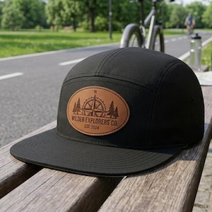 May include: Black five-panel cap with a flat brim. A brown leather patch features a compass design, trees, and the text "Wilder Explorers Co. EST. 2024". The cap is sitting on a wooden bench outdoors.