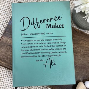 May include: A teal-colored journal with the words "Difference Maker" in black script. The journal includes a definition and the name "Ali" in cursive. The journal is on a wooden surface.