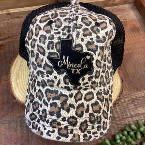 May include: A trucker hat with a leopard print pattern in shades of brown and tan. The hat features a black mesh back and a black patch with the text "Mineola TX" in a script font, with a Texas outline.