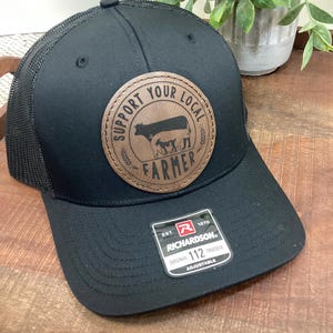 May include: Black trucker hat with a brown leather patch that says "Support Your Local Farmer" with a silhouette of a cow and two calves. The hat is adjustable and has the Richardson 112 logo on the side.
