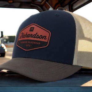 May include: A navy blue and tan trucker hat with a Richardson patch. The patch is a red-orange hexagon with the brand name and "PREMIUM HEADWEAR 1970" printed on it. The hat has a mesh back and a curved brim.