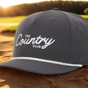 May include: A gray baseball cap with the text "The Country Club" in white script. The cap has a white rope detail along the brim and is set against a golf course background.
