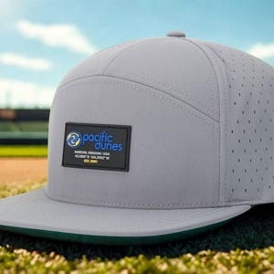 May include: A gray baseball cap with a flat brim and a green underside. The cap features a perforated design on the sides and a rectangular patch with the text "pacific dunes". The cap is sitting on a baseball field.