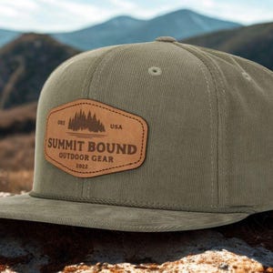 May include: Olive green corduroy baseball cap with a flat brim. A brown leather patch features a mountain and tree graphic with the text "SUMMIT BOUND OUTDOOR GEAR 2022" and "USA". The cap is designed for outdoor activities.