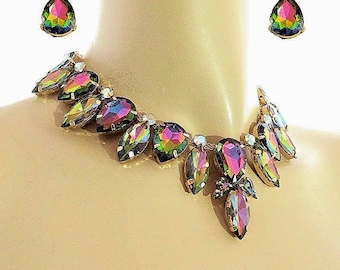 May include: A necklace and earring set featuring iridescent, multi-colored, pear-shaped crystals set in gold-toned metal. The necklace consists of a series of these faceted stones arranged in a collar style, with matching teardrop earrings.