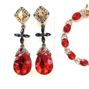 May include: A set of gold-toned jewelry featuring a pair of drop earrings and a bracelet. The earrings have a square top with a clear stone, a blue cross design, and a large red teardrop-shaped stone. The bracelet has red oval stones.